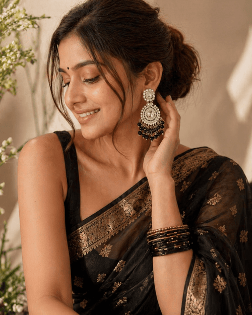 Woman wearing a black saree with gold patterns and large earrings, posing indoors.