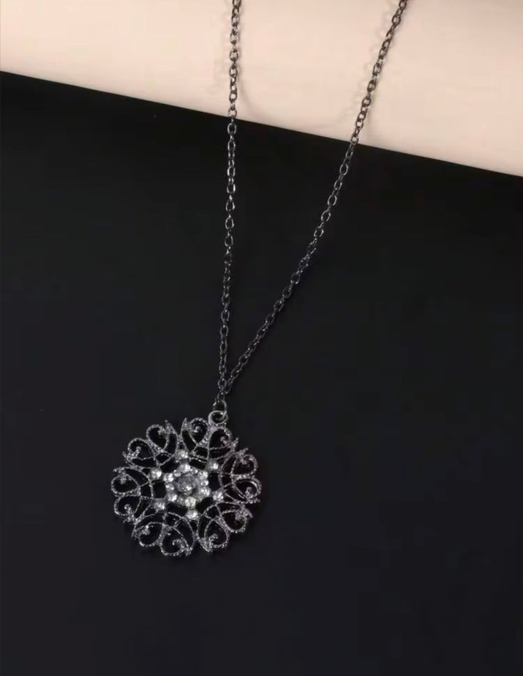 A silver filigree pendant necklace with a floral center, displayed on a black surface with a cream-colored background at the top.