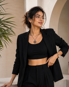 A woman wearing a black tailored blazer over a black crop top and black trousers, standing in a softly lit architectural setting with beige walls and a plant at the left edge.
