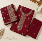 Three folded pieces of deep maroon unstitched dress material with ornate beige and gold floral embroidery, arranged on a light cream background with decorative props.