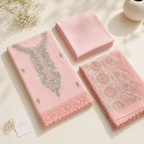 Three folded pink fabric garments or textiles displayed on a light cream surface with soft shadows. The left item is a pink embroidered piece with a scalloped lace hem and gray floral embroidery; the center item is a solid pink folded textile; the right item is a pink patterned textile with circular motifs and lace edges.