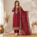 A woman models a deep maroon three-piece salwar kameez suit with intricate gold embroidery, matching trousers, and a coordinating dupatta, set against a warm, sunlit room with decorative vases.