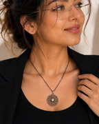 A close-up of a woman wearing a round, silver-toned pendant necklace with a central clear stone and textured outer details, styled with a black top and blazer.